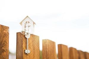 Small figure of white wooden house on fence with key to housing against the background of cottages. Building, design, project, moving to new house, mortgage, rent and purchase real estate. Copy space