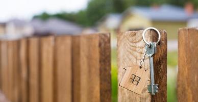 Wooden pendant of a house and key. Background of fence and cottage. Dream of home, building, design, delivery of the project, moving to a new house, mortgage, rent and purchase real estate. Copy space photo