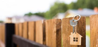 Wooden pendant of a house and key. Background of fence and cottage. Dream of home, building, design, delivery of the project, moving to a new house, mortgage, rent and purchase real estate. Copy space photo