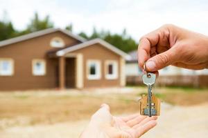 Hand with a key and a wooden key ring-house. Background of fence and cottage. Building, project, moving to a new home, mortgage, rent and purchase real estate. To open the door. Copy space photo