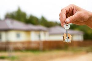 Hand with a key and a wooden key ring-house. Background of fence and cottage. Building, project, moving to a new home, mortgage, rent and purchase real estate. To open the door. Copy space photo