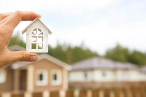 Model of white wooden house in hand against the background of cottages in the village. Dream of home, building, design, moving to a new house, mortgage, rent and purchase real estate. Copy space photo