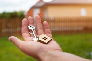 Wooden pendant of a house and key. Background of fence and cottage. Dream of home, building, design, delivery of the project, moving to a new house, mortgage, rent and purchase real estate. Copy space photo