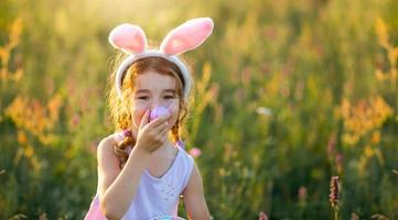 Cute funny girl with painted Easter eggs in spring in nature in a field with golden sunlight and flowers. Easter holiday, Easter bunny with ears, colorful eggs in a basket. Lifestyle photo