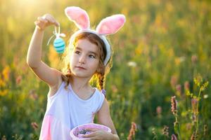 Cute funny girl with painted Easter eggs in spring in nature in a field with golden sunlight and flowers. Easter holiday, Easter bunny with ears, colorful eggs in a basket. Lifestyle