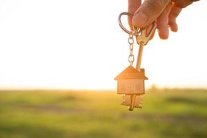 House key with keychain in hand. Background of sky, sunlight and field. Dream of home, building a cottage in countryside, plan and project design, farm, moving to a new home, accommodation. Copy space photo
