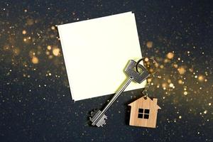 Keychain in the shape of wooden house with key on a black background with a square sheet for notes. Building, design, project, moving to new house, mortgage, rent and purchase real estate. Copy space photo