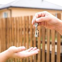 Hand with a key and a wooden key ring-house. Background of fence and cottage. Building, project, moving to a new home, mortgage, rent and purchase real estate. To open the door. Copy space photo
