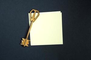 A golden key on a piece of note paper. Wish list, problem solution, idea, decoding, business concept on a black background with a square sheet. Project, rent, booking, household chores. Copy space photo