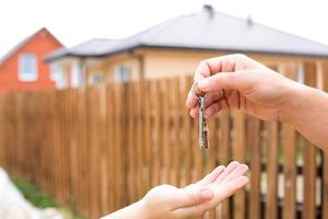 Hand with a key and a wooden key ring-house. Background of fence and cottage. Building, project, moving to a new home, mortgage, rent and purchase real estate. To open the door. Copy space photo