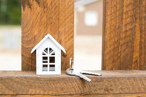 Small figure of white wooden house on fence with key to housing against the background of cottages. Building, design, project, moving to new house, mortgage, rent and purchase real estate. Copy space photo