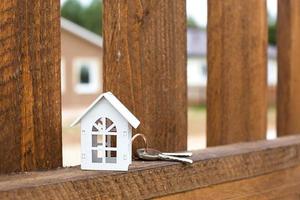 Small figure of white wooden house on fence with key to housing against the background of cottages. Building, design, project, moving to new house, mortgage, rent and purchase real estate. Copy space