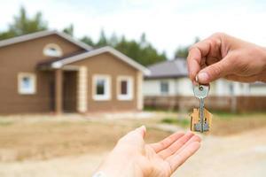 Hand with a key and a wooden key ring-house. Background of fence and cottage. Building, project, moving to a new home, mortgage, rent and purchase real estate. To open the door. Copy space photo