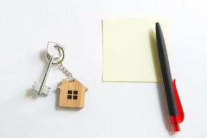 Keychain in the shape of wooden house with key on a white background with a square sheet for notes. Building, design, project, moving to new house, mortgage, rent and purchase real estate. Copy space photo