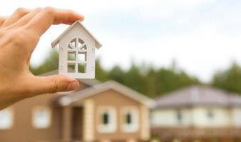 modelo de casa de madera blanca en la mano contra el fondo de las cabañas en el pueblo. sueño de casa, construcción, diseño, mudanza a una nueva casa, hipoteca, alquiler y compra de bienes raíces. copie el espacio foto