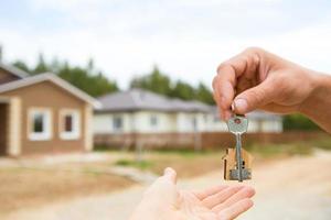 Hand with a key and a wooden key ring-house. Background of fence and cottage. Building, project, moving to a new home, mortgage, rent and purchase real estate. To open the door. Copy space photo