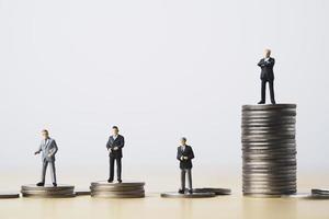 figura en miniatura de cuatro hombres de negocios parados en diferentes monedas altas y bajas apiladas para la posición y el concepto exitoso de ingresos de inversión empresarial. foto