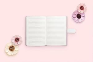 Notepad surrounded by flowers on pink surface. Blank papers for text. Flat lay, top view composition photo