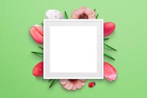 Blank picture frame surrounded by Easter eggs and flowers on green surface. Top view, flat lay composition photo