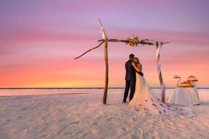 The bride and groom under wedding arch on beach. Romantic wedding background. Amazing beach sunset, infinity sea view, romantic clouds sky. Idyllic destination honeymoon