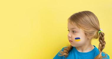 una chica con una bandera ucraniana pintada de amarillo y azul en las mejillas. copie el espacio sobre fondo amarillo. foto