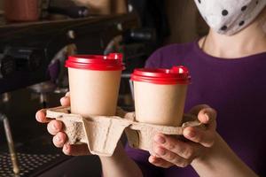 tazas de cartón simuladas con capuchino en un puesto de comida para llevar en manos de una mujer con máscara facial. café para llevar concepto foto