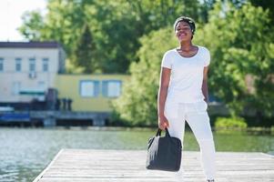 retrato de una elegante chica afroamericana, vestida con ropa blanca, con una bolsa en la mano contra el muelle del lago. moda callejera de jóvenes negros.