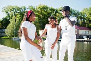 Three stylish, cheerful and trendy african american friends, wear on white clothes. Street fashion of young black people. Black man with two african girls. photo