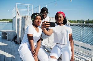 Three stylish african american friends, wear on white clothes at pier on beach making selfie. Street fashion of young black people. Black man with two african girls. photo