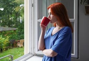 young woman looking out of window while drinking from coffee cup