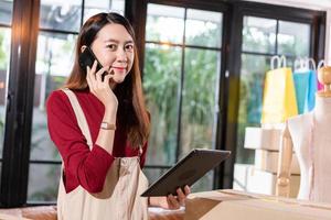 Young Asian shop owner woman calling to customer and checking stock on digital tablet at home. SME small business entrepreneur prepare for shipping product, online store e-commerce or service support photo