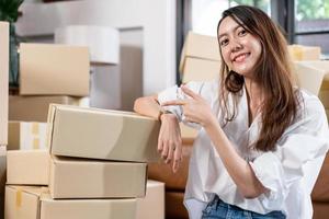 SME small business Asian female entrepreneur or shop owner happy hand pointing to parcel boxes before delivery to customer. freelancer, online shopping, startup and e-commerce concept