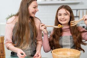 feliz madre e hija disfrutan cocinando ensalada casera en la cocina. chica sonriente preparando verduras. actividad infantil con los padres en casa. foto