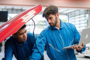 Caucasian automobile mechanic man using laptop computer diagnostic and repairing car while his colleague checking in radiator bonnet at garage automotive, motor technician maintenance after service photo