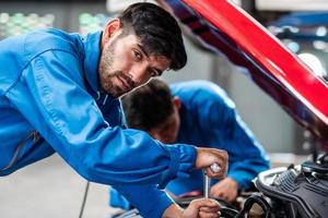 Mecánico de automóviles caucásico comprobando el estado de la pieza rota del daño del automóvil, diagnosticando y reparando el vehículo en el garaje automotriz, mantenimiento técnico del motor después del concepto de servicio foto