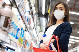 Beautiful woman wearing medical face mask and rubber glove with grocery trolley picking up daily milk on product shelf. shopping at supermarket in new normal lifestyle concept during Coronavirus. photo