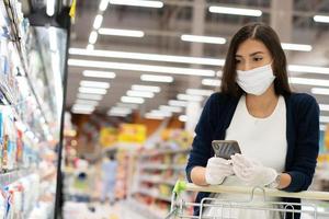 Woman wearing protective mask and rubber glove checking shopping list on mobile phone to buy stuff at grocery store. new normal lifestyle during Coronavirus pandemic photo