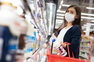 hermosa mujer con mascarilla médica y guante de goma con carro de supermercado recogiendo leche diaria en el estante del producto. compras en el supermercado en un nuevo concepto de estilo de vida normal durante el coronavirus. foto