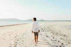 un hombre con camisa blanca caminando en una playa de arena blanca mientras mira al mar foto