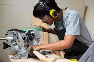 un joven carpintero que trabaja en su mesa de taller usando una sierra circular y usando equipo de seguridad foto