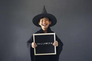 lindo niño asiático celebrando halloween vistiendo un disfraz de bruja y sosteniendo accesorios de decoración de halloween foto
