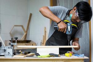 un joven carpintero que trabaja en su mesa de taller usando una máquina perforadora y usando equipo de seguridad