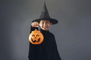 lindo niño asiático celebrando halloween vistiendo un disfraz de bruja y sosteniendo accesorios de decoración de halloween foto