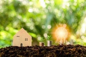 A bright light bulb and simulated wooden house on the mound. The concept for realty, economy, finance, coins. Daytime, nature, clear air, the light of shine in the morning. Blurred, bokeh background photo
