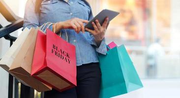 mujer usando tableta y sosteniendo una bolsa de compras de viernes negro mientras está de pie en las escaleras con el fondo del centro comercial foto