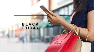 viernes negro, mujer usando un teléfono inteligente y sosteniendo una bolsa de compras mientras está de pie en el fondo del centro comercial foto