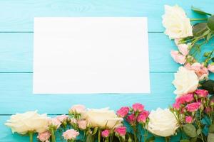 Frame of colorful roses on blue wooden background with paper card and copy space. Top view and selective focus. Mock up photo