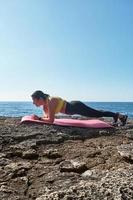 Latin woman, middle-aged, with yellow top, black leggings, training, doing physical exercises, plank, abs, climber's step, burning calories, keeping fit, outdoors by the sea,blue sky, photo