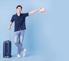 Image of young Asian man holding suitcase on blue background, travel concept photo