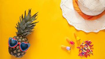 Summer composition. Pineapple in sunglasses, hat, beads and nail polishes on a yellow background with copy space. Vacation concept. Flat lay.